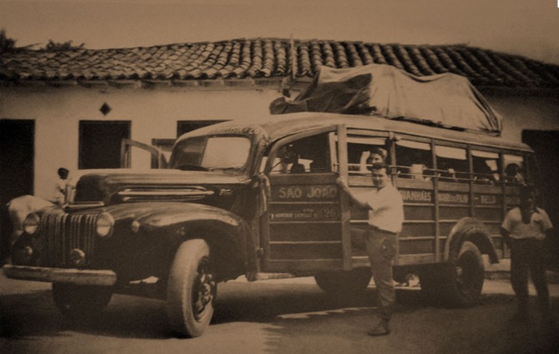 Fotografia em preto e branco de um caminhão adaptado para transporte de passageiros, com carroceria de madeira e o nome São Jorge na lateral. Dois homens posam ao lado do veículo.